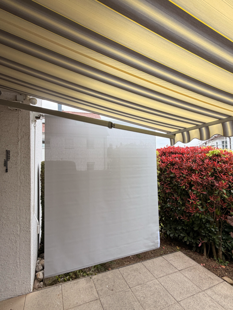 Seitenbeschattung, Skuggi neben einer Markise Ein Wandbereich mit einem weißen Sichtschutz und roten Blumen im Hintergrund.